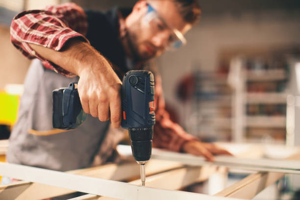 Carpenter using a cordless power drill to assemble a wooden frame in a workshop, wearing safety goggles, signifying the most essential tools