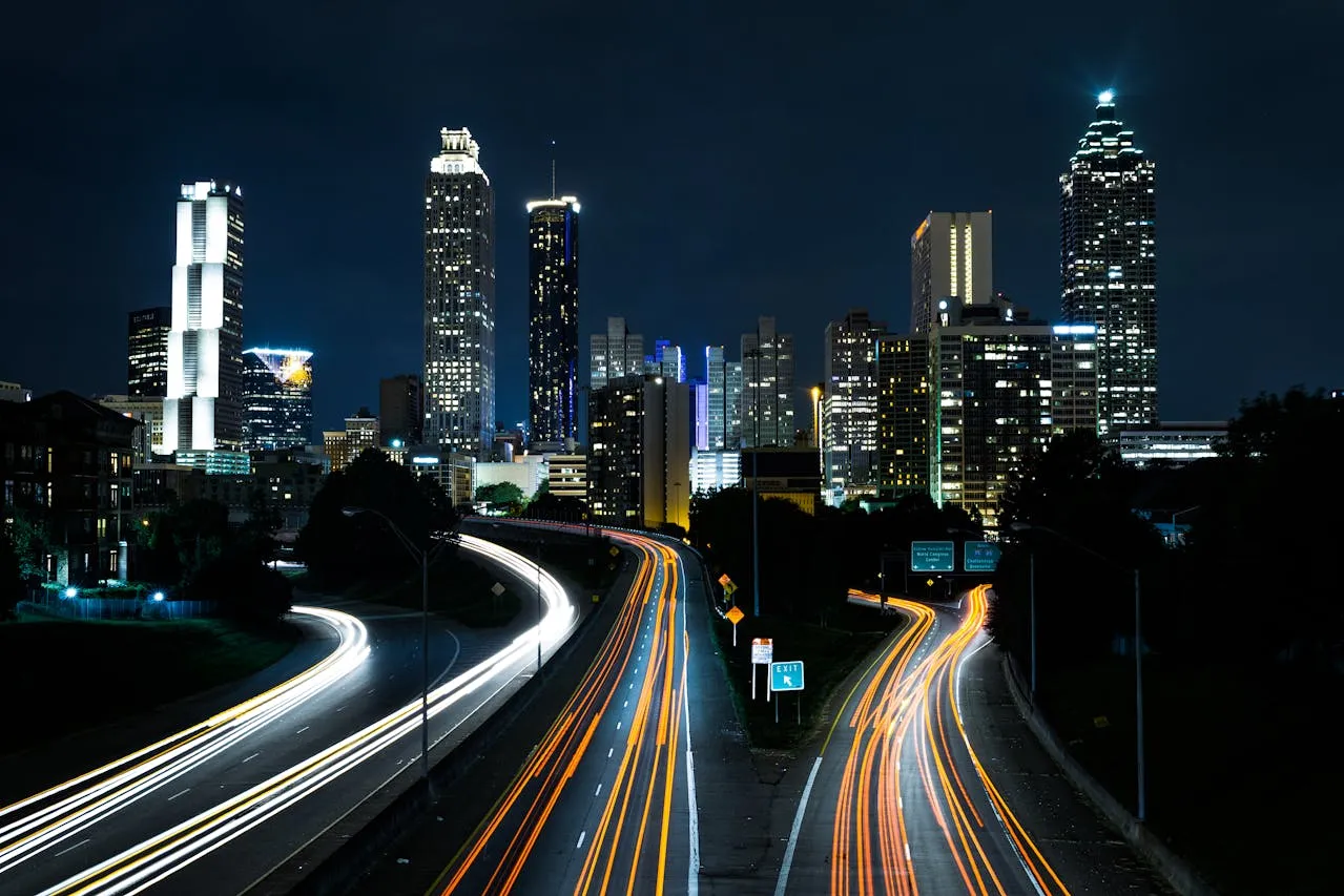 A nighttime cityscape featuring illuminated skyscrapers and a highway with long-exposure light trails from moving vehicles.