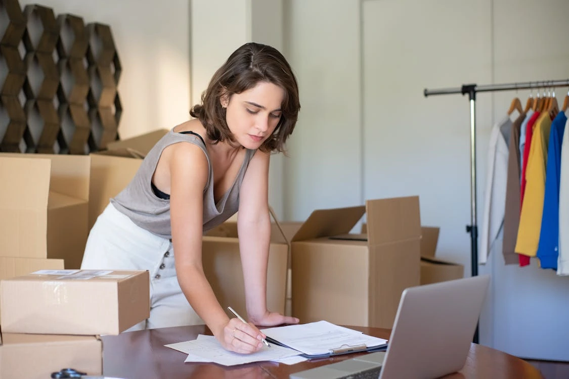 A young woman leaning over a desk while reviewing documents in a workspace surrounded by cardboard boxes, clothes on a rack, and a laptop, representing a small ecommerce business owner managing orders.
