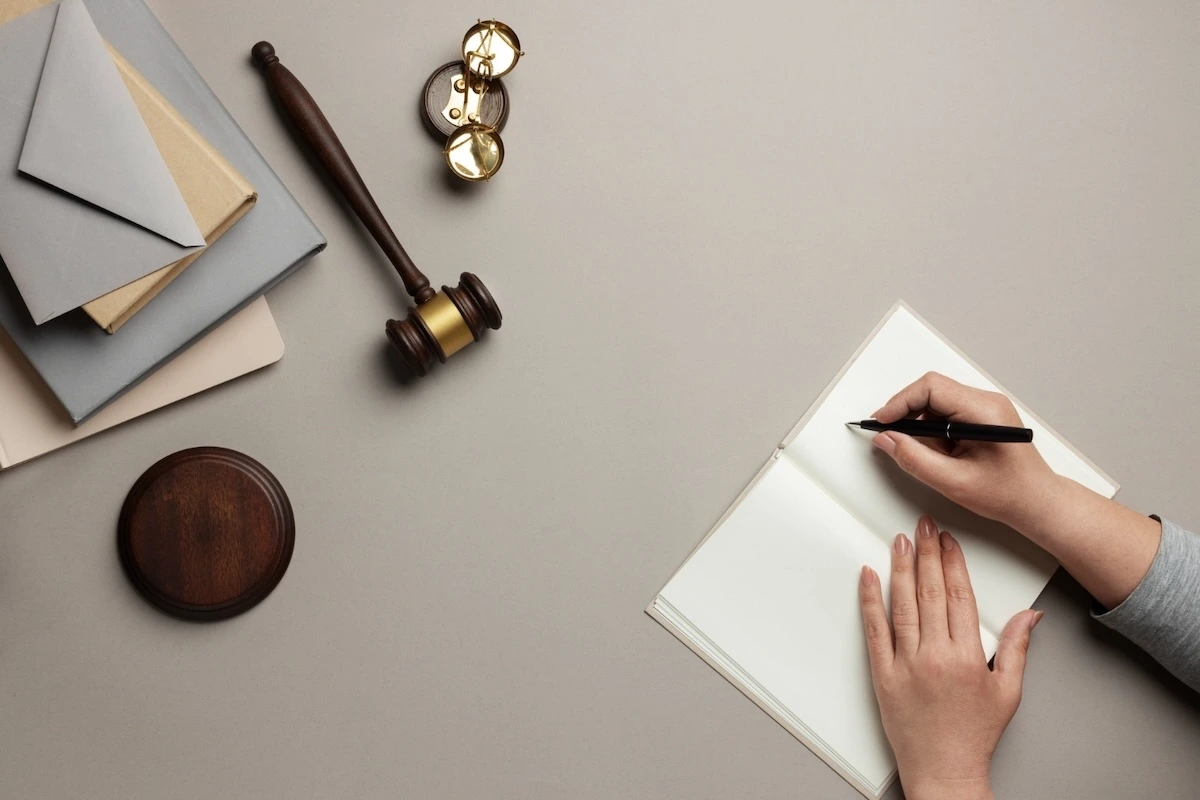 A flat lay image showing a legal-themed workspace with a gavel, round wooden sound block, a pair of golden scales, two closed folders with envelopes, and a person's hands writing in a blank notebook with a black pen.
