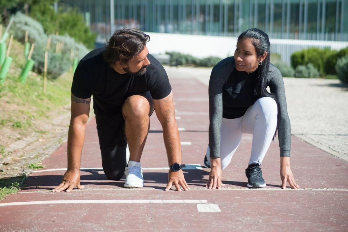 Two athletes, a man and a woman, are in a sprint start position on an outdoor running track, looking at each other and smiling, suggesting a friendly competition or training session.