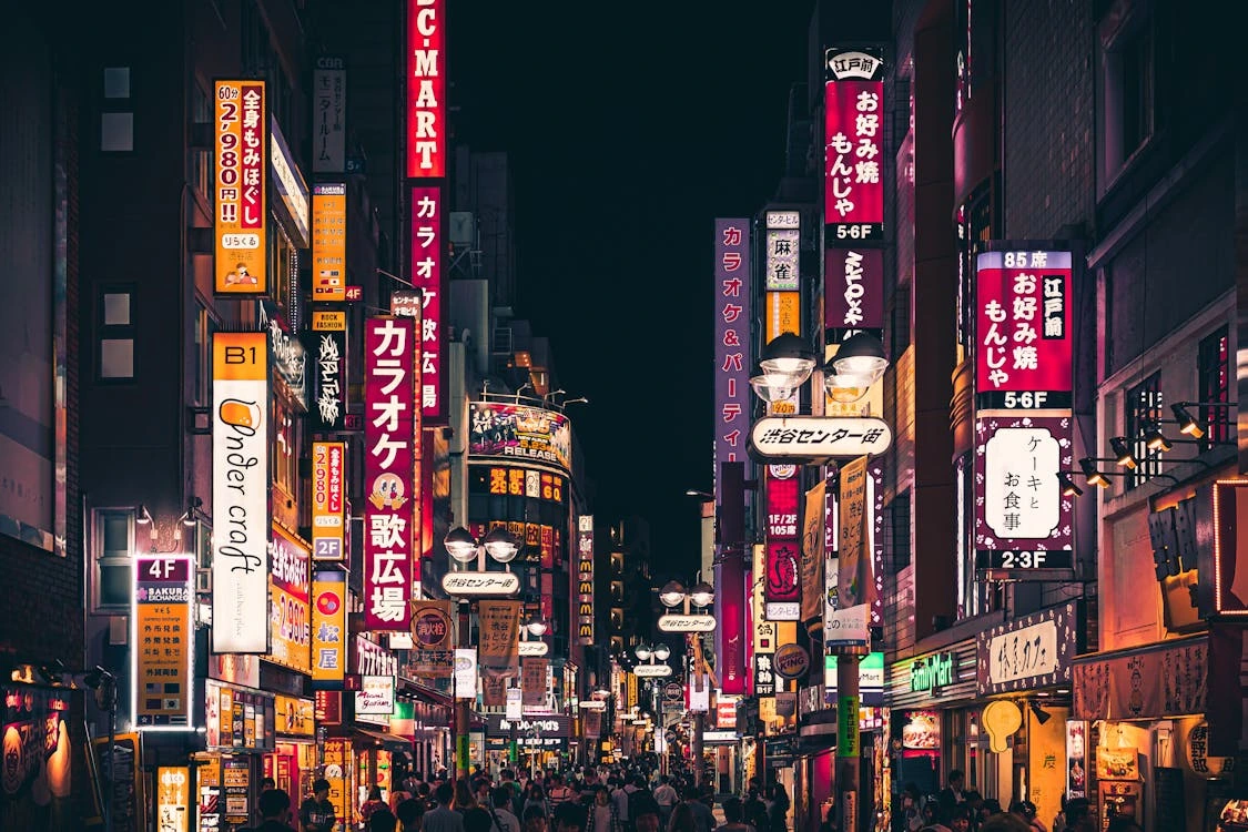 A vibrant street scene at night in a bustling Japanese city, illuminated by numerous colorful neon signs in Japanese characters, advertising karaoke bars, restaurants, and shops. The street is crowded with people walking under the bright lights and overhead lamps.
