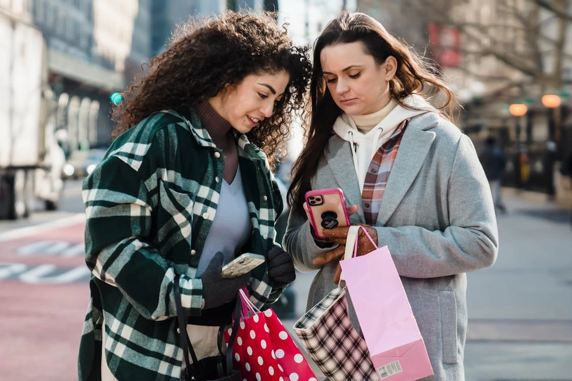 "Two women standing outdoors on a city street during daytime, holding shopping bags and looking at a smartphone together. One woman wears a green plaid coat and holds a white phone, while the other wears a grey coat with a layered hoodie and flannel shirt, holding a pink phone. They appear to be engaged in a conversation or checking something online while shopping."