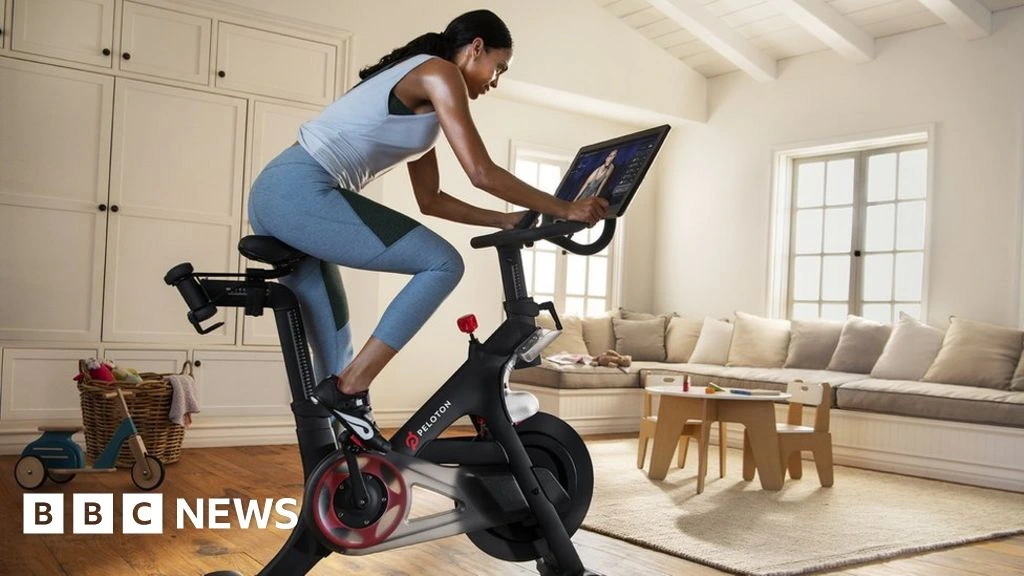 A woman rides a Peloton exercise bike in a stylish living room, following a virtual fitness class on the screen, with BBC News branding in the corner.