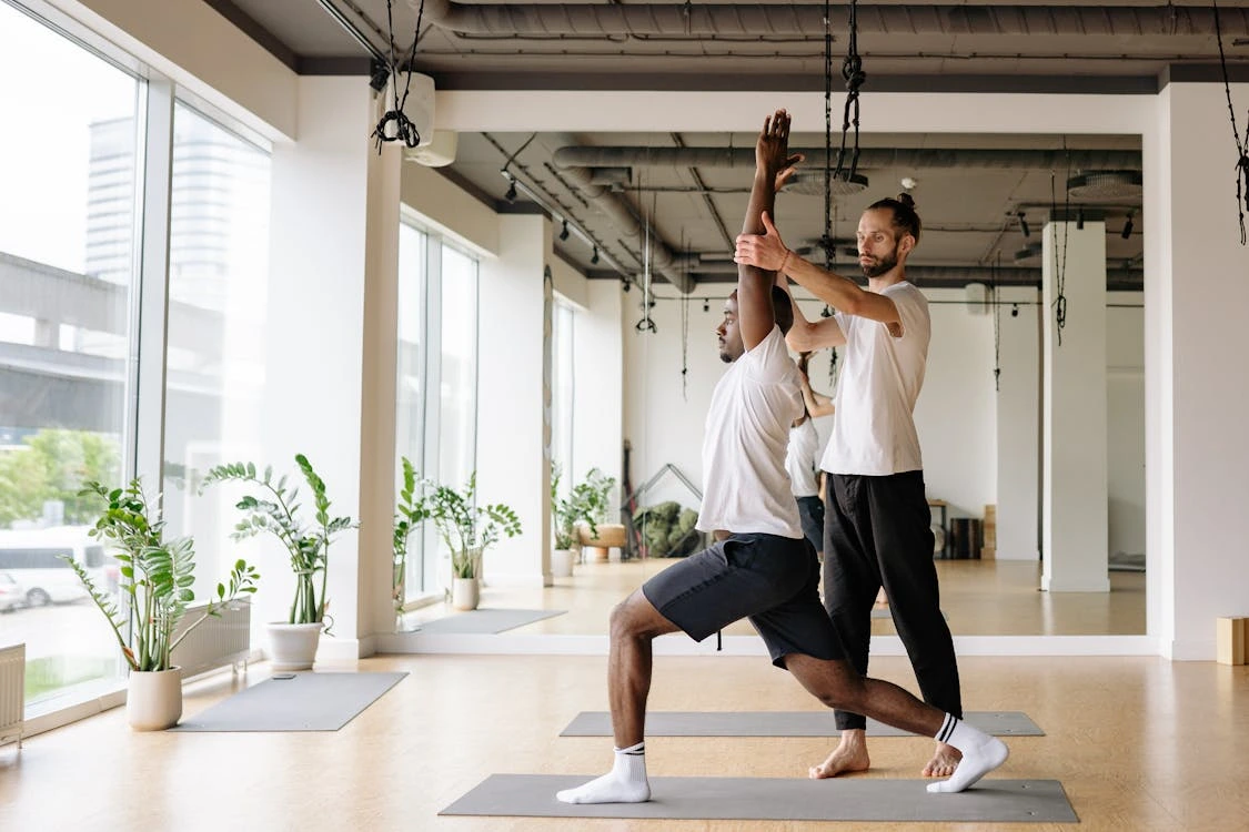 A yoga instructor assists a student in a lunge pose at a bright, modern studio with large windows and potted plants.