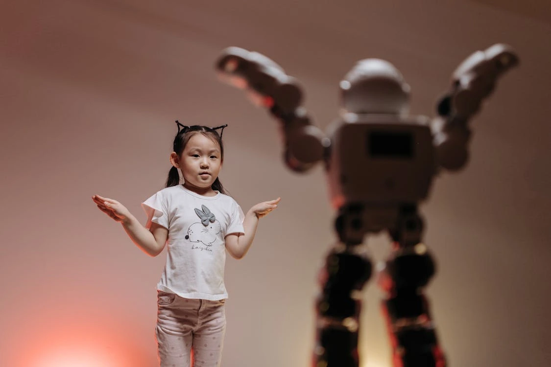 A young girl wearing cat ear headbands and a white t-shirt with a cartoon character stands with her arms raised slightly to the sides, mimicking or interacting with a humanoid robot. The robot, larger and in the foreground, has its arms raised as well. The background is softly lit with a warm, neutral tone, creating a playful and curious atmosphere.
