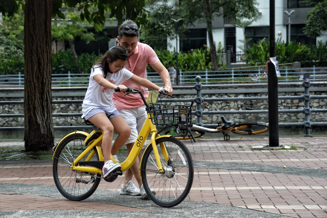 “A man in a pink shirt helping a young girl ride a yellow Ofo bicycle in a park, with another parked bike lying on the ground nearby.”