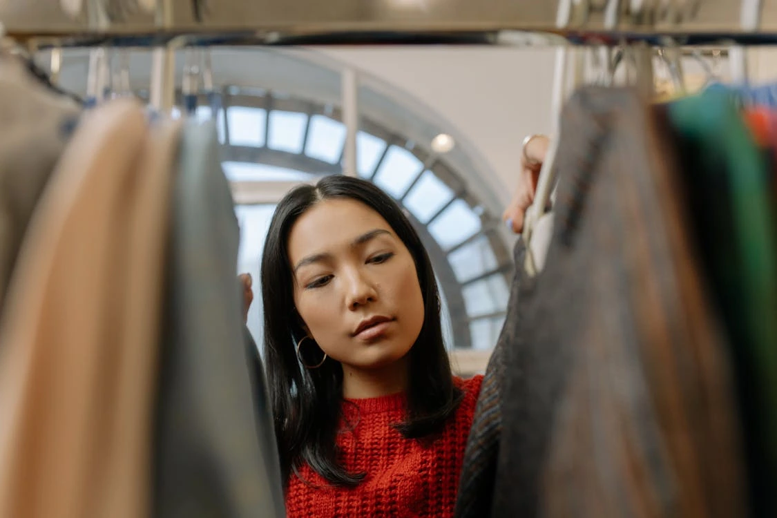 A woman in a red sweater thoughtfully browsing through clothes on a rack inside a well-lit store with arched windows in the background.