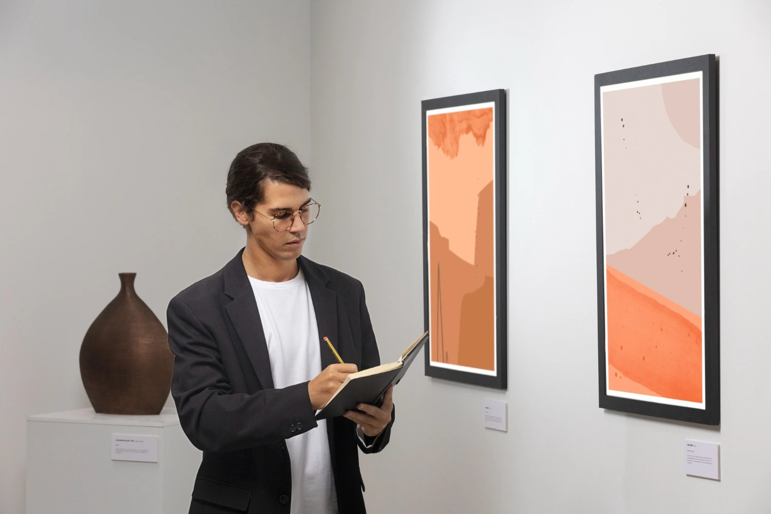 A man with glasses and a black blazer takes notes in a notebook while standing in front of two abstract orange-toned paintings at an art gallery. A ceramic vase is visible on a pedestal behind him.