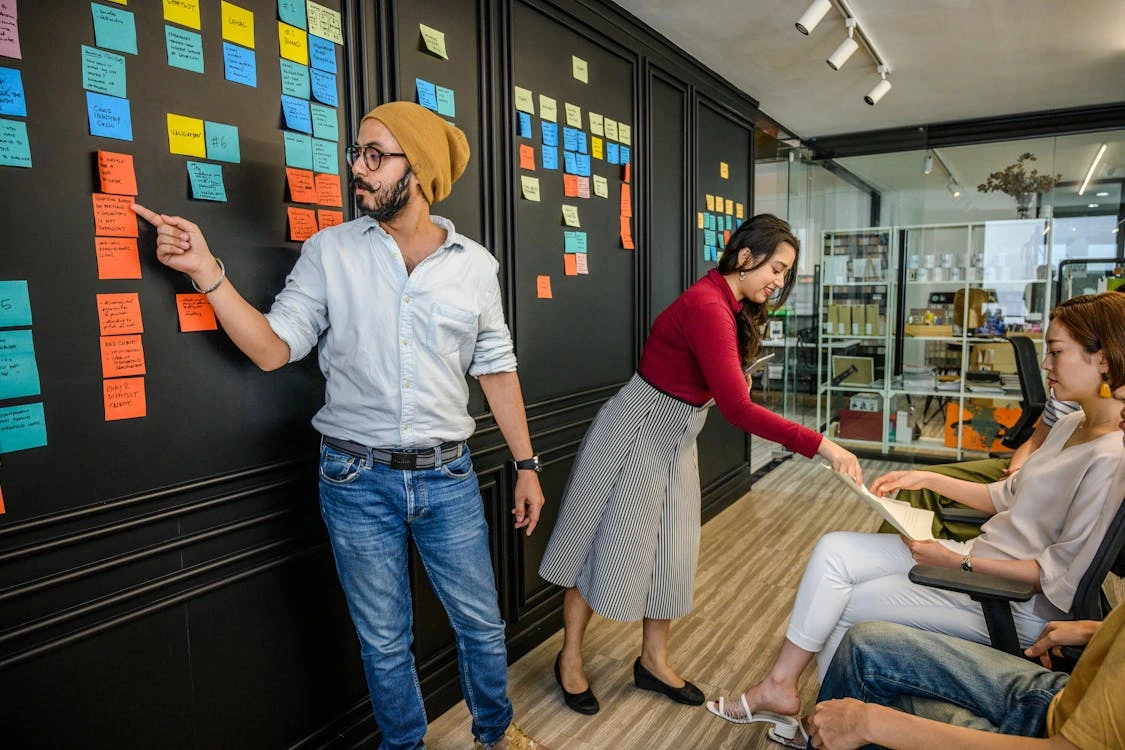 A group of colleagues in a modern office space collaborating during a planning session. One man in a beanie is pointing at colorful sticky notes on a black wall, while a woman in a red top and striped skirt hands a document to a seated woman. Another person is seated and listening. The wall is filled with organized sticky notes for brainstorming or task tracking.