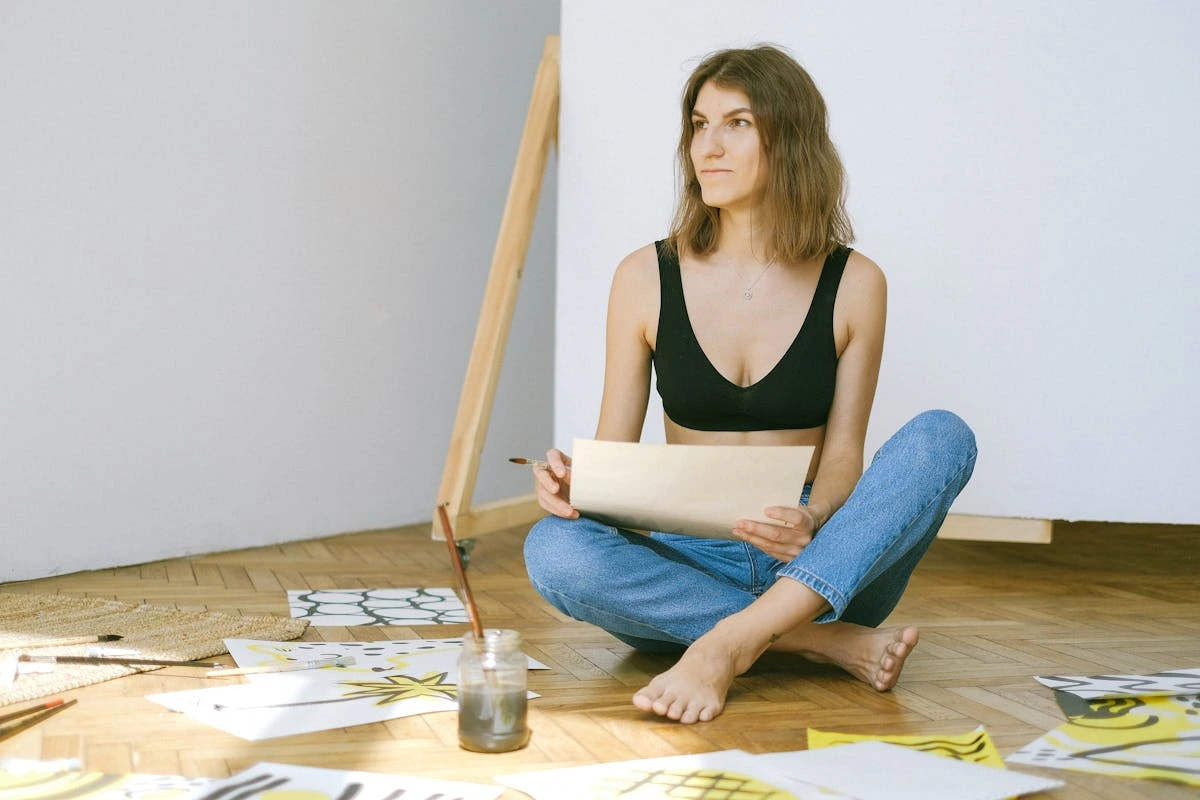 A young woman sitting on the floor in a sunlit room surrounded by abstract art pieces and painting supplies, holding a sheet of paper with a thoughtful expression.