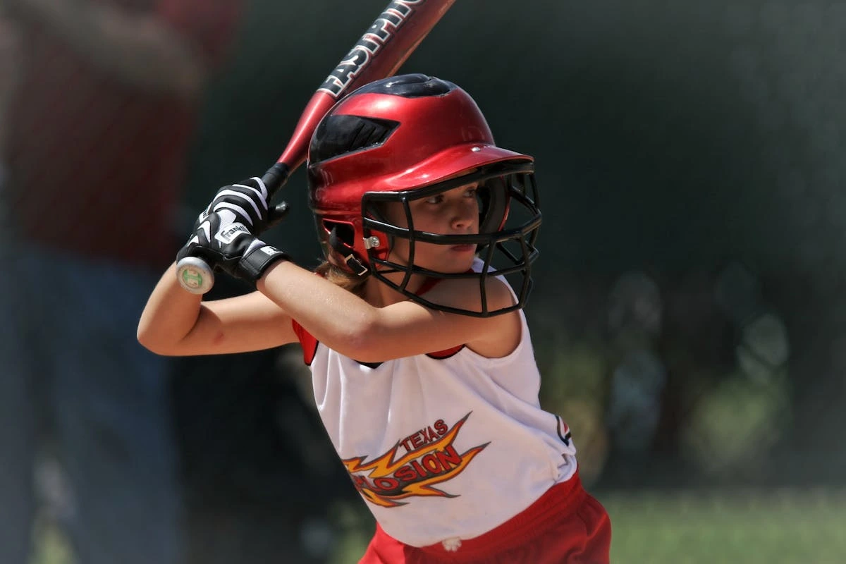 A young girl wearing a red helmet and white jersey with "Texas Explosion" logo prepares to swing a baseball bat during a game. She is focused, gripping the bat with both hands, wearing black-and-white gloves, and standing in a batter’s stance on a sunny day.