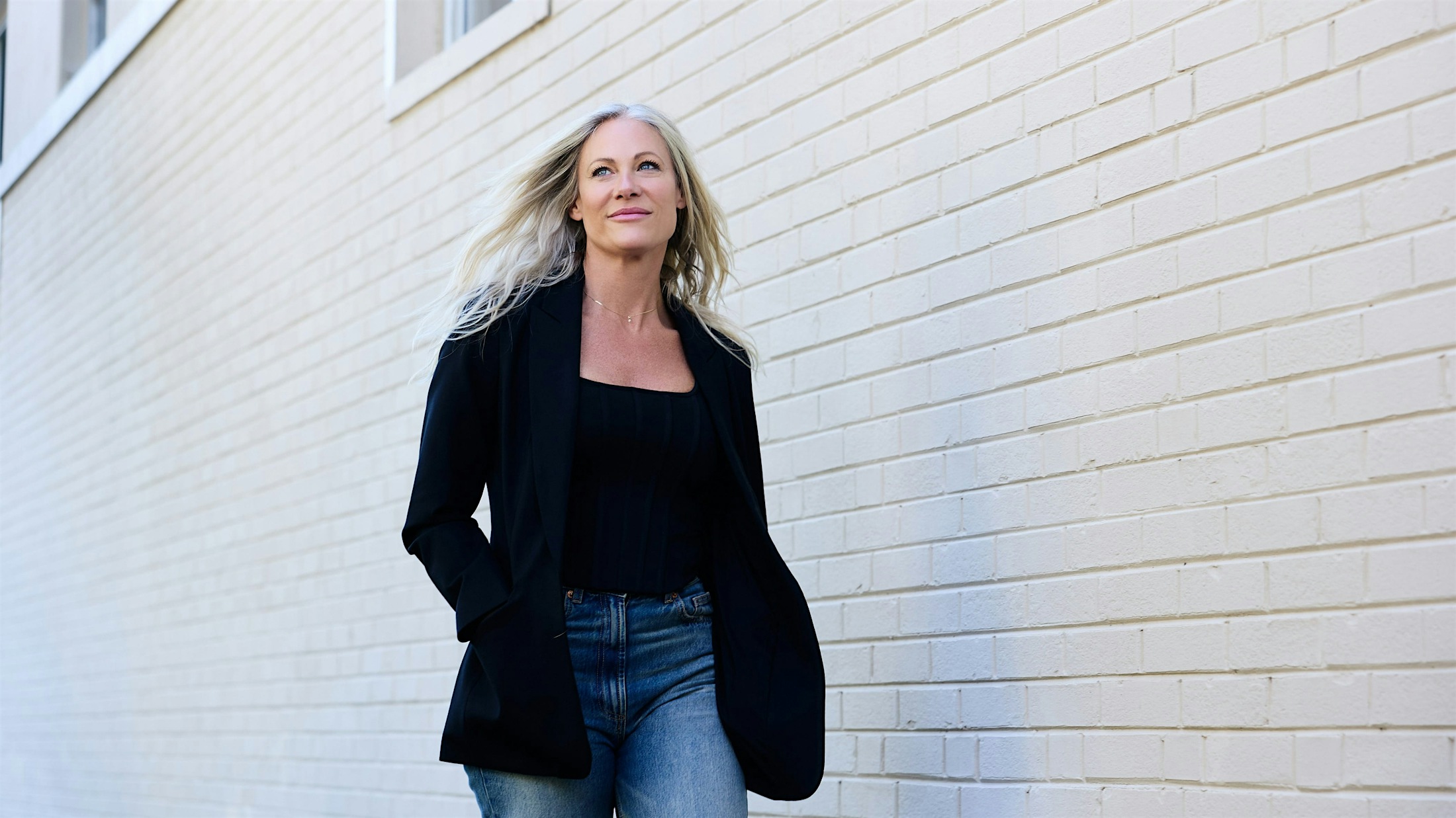 woman with long blond hair walks down the side of a brick building