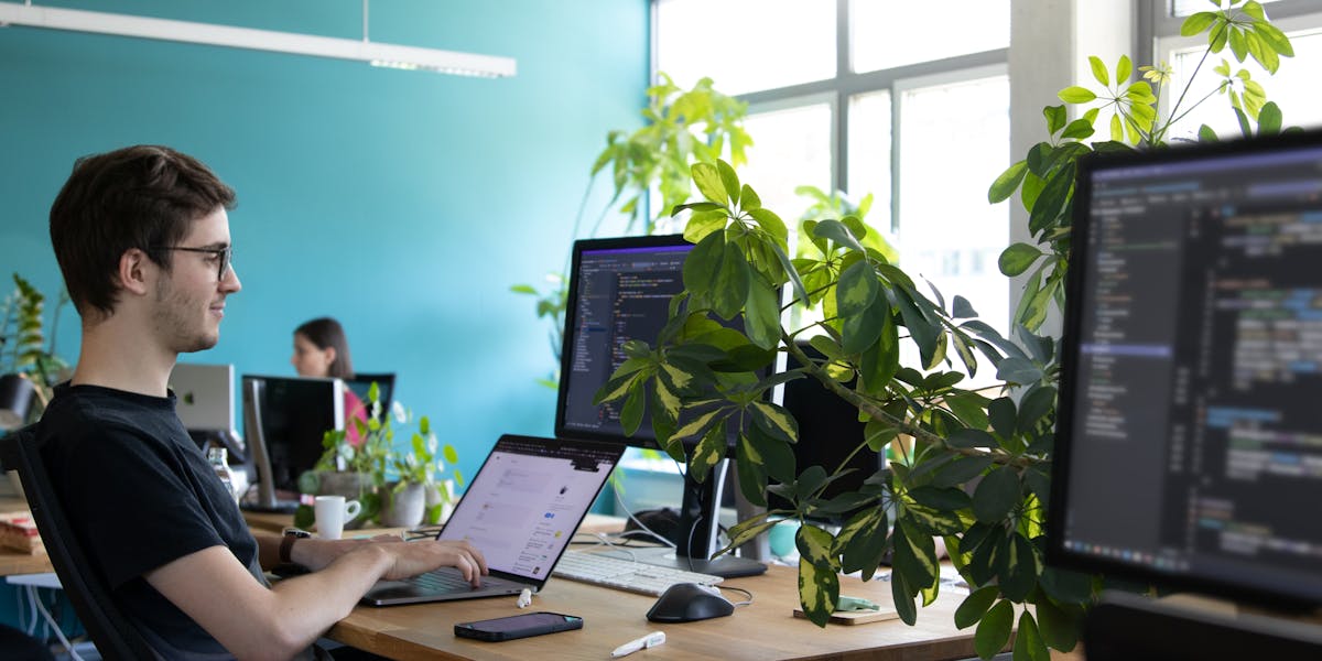 Web developer vibecoding in the Peerigon office Entwickler arbeitet an seinem Schreibtisch mit Laptop und Monitor in einem modernen Büro mit grüner Wand und Pflanzen.