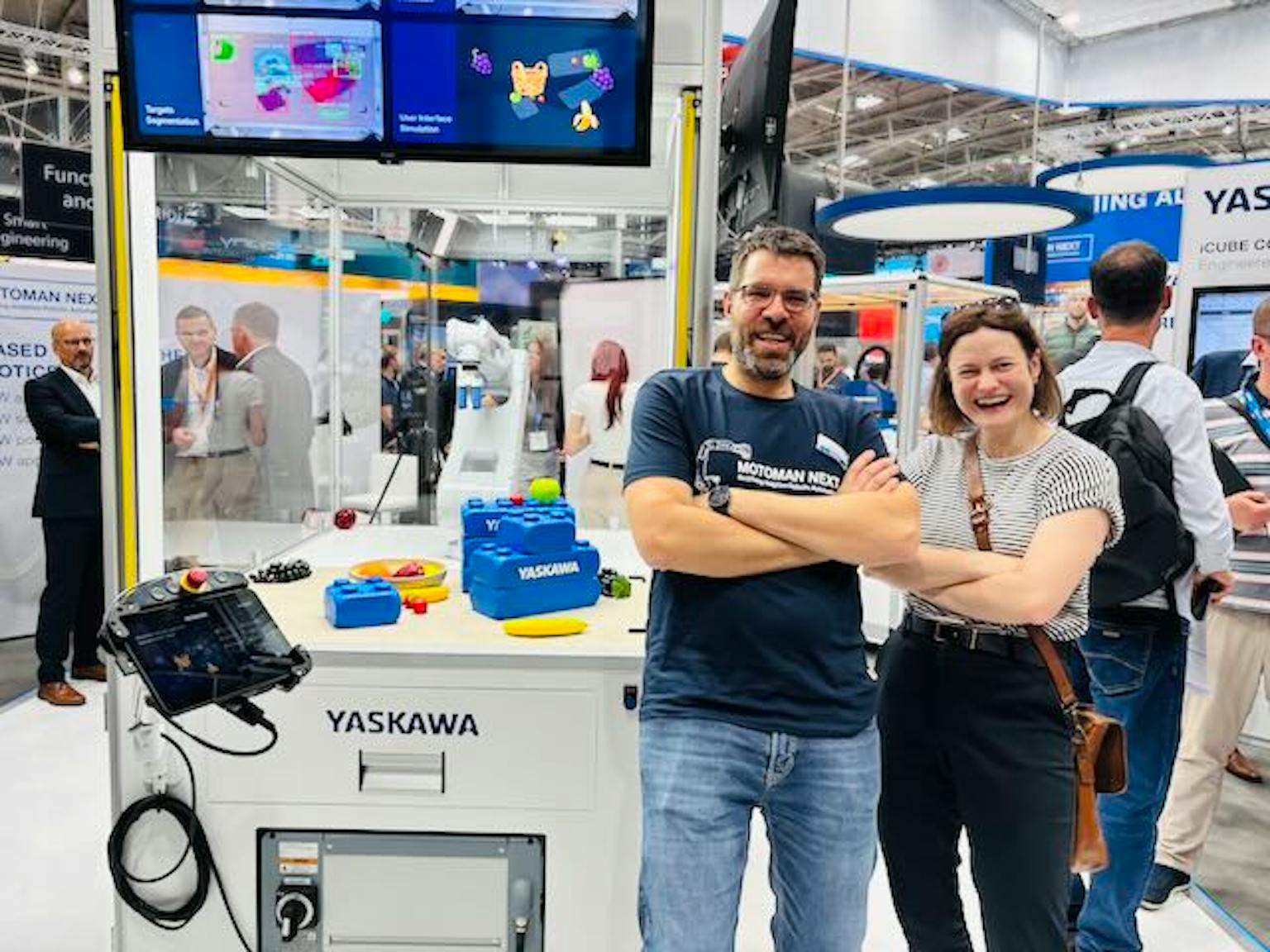 YASKAWA Messestand mit Mitarbeitern Zwei Personen posieren vor einem YASKAWA-Messestand mit blauen Produktmodellen und Bildschirmen im Hintergrund