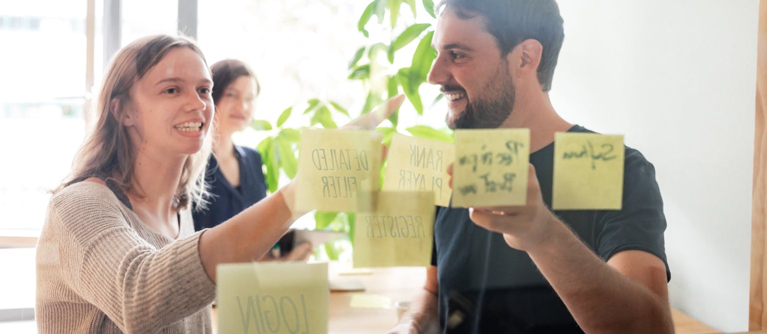 Two people place sticky notes on a glass wall during a workshop or brainstorming session in an office setting.