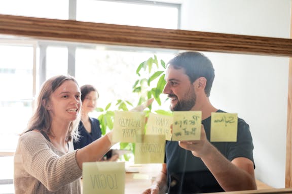 Two employees discussing sticky notes on a glass wall during a workshop or brainstorming session.