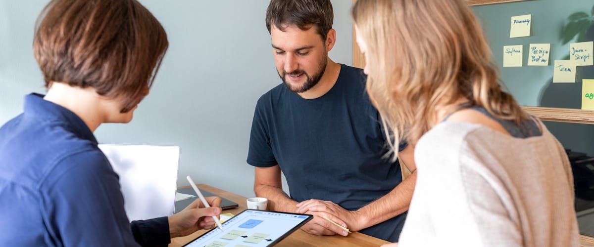 Design Team Collaboration Meeting Three people collaborating at a table, reviewing design mockups on a tablet with notes and coffee.