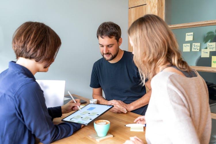 Three people collaborating at a table, reviewing design mockups on a tablet with notes and coffee.