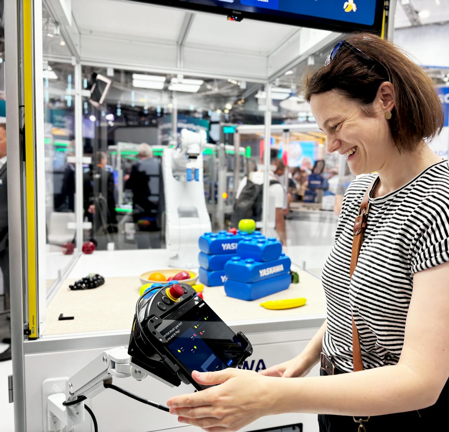 Yaskawa HMI in der Praxis Frau hält ein schwarzes Yaskawa-Steuergerät in den Händen und lächelt, während sie es in einem modernen Fabrikationslabor betrachtet