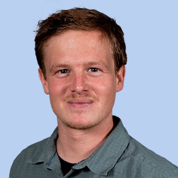 Team Portrait Michael J. Portrait photo of a man with brown hair and mustache wearing a gray shirt against a blue background