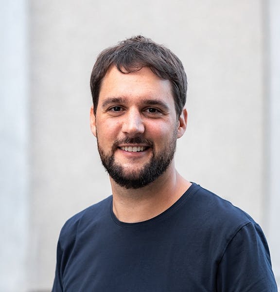 Portrait eines lächelnden Mannes mit dunklem Bart, schwarzem T-Shirt vor hellem Hintergrund