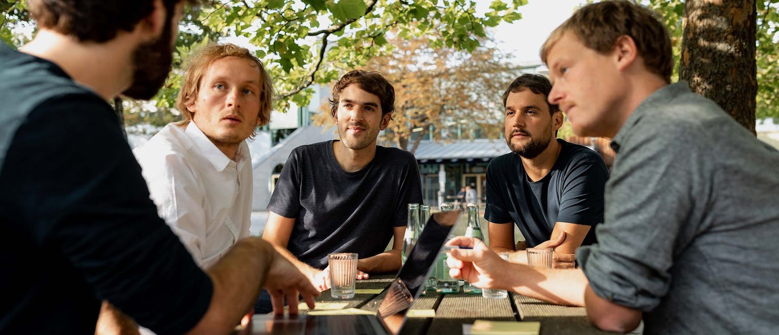 Team Discussion Outdoors Five men sit together at an outdoor table beneath trees, engaged in an animated discussion.