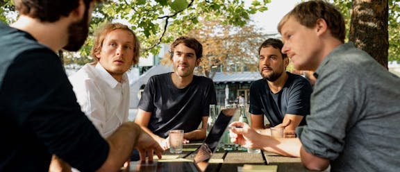 Team Discussion Outdoors Five men sit together at an outdoor table beneath trees, engaged in an animated discussion.