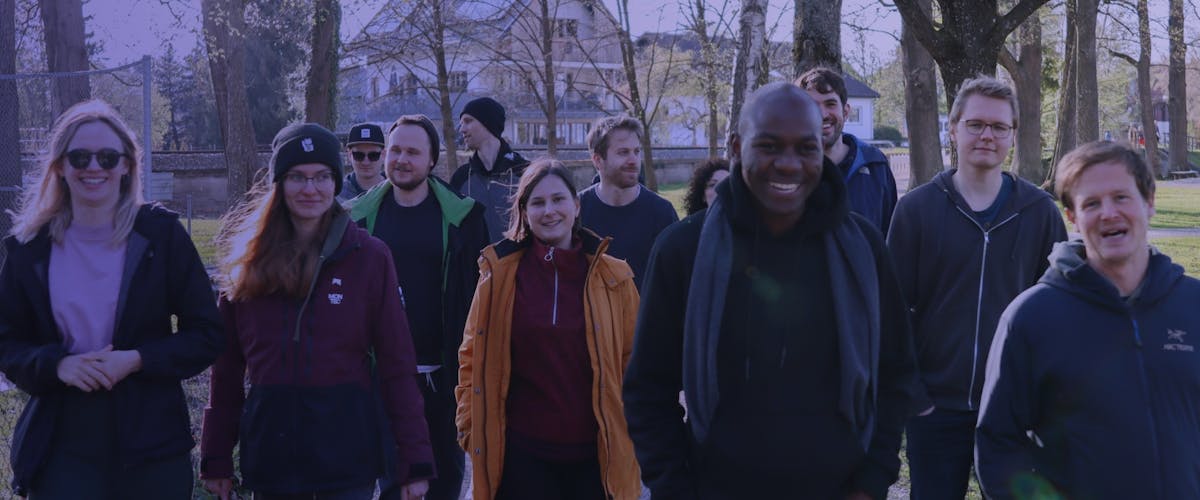Peerigon Team Gruppe von etwa zehn Personen steht zusammen in einem Park vor Bäumen und lächelt in die Kamera