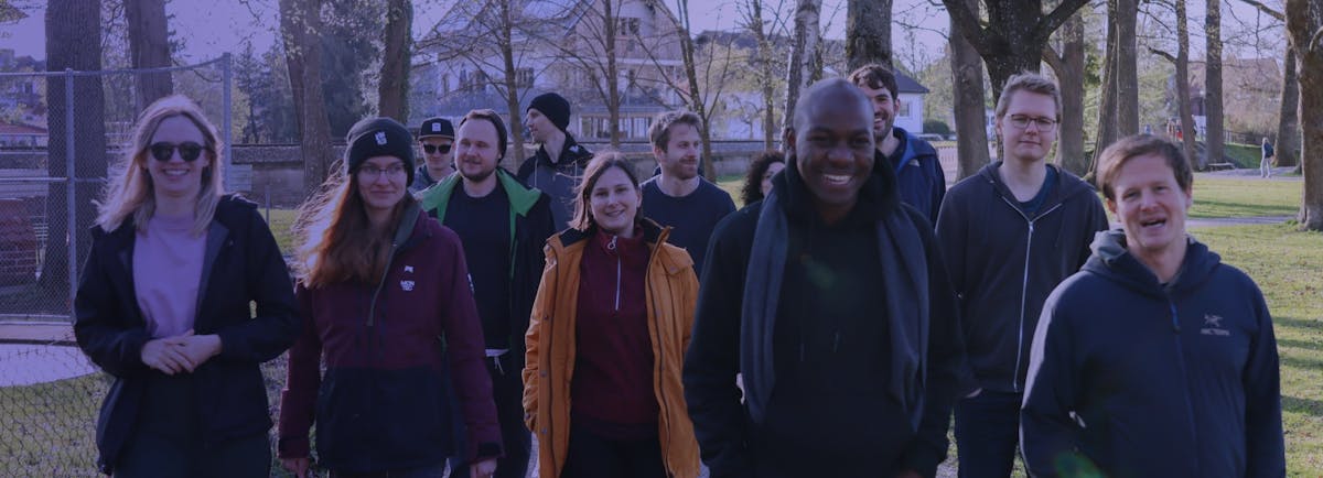 Gruppe von etwa zehn Personen steht zusammen in einem Park vor Bäumen und lächelt in die Kamera