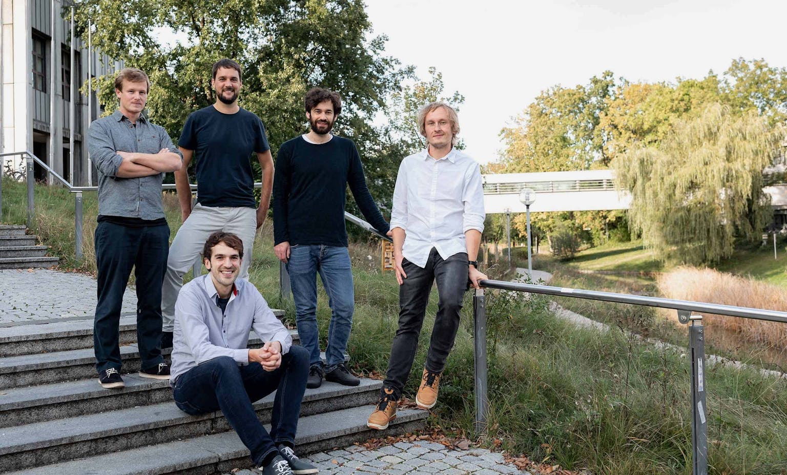 Five men pose together on outdoor steps next to a building overlooking a park and river.