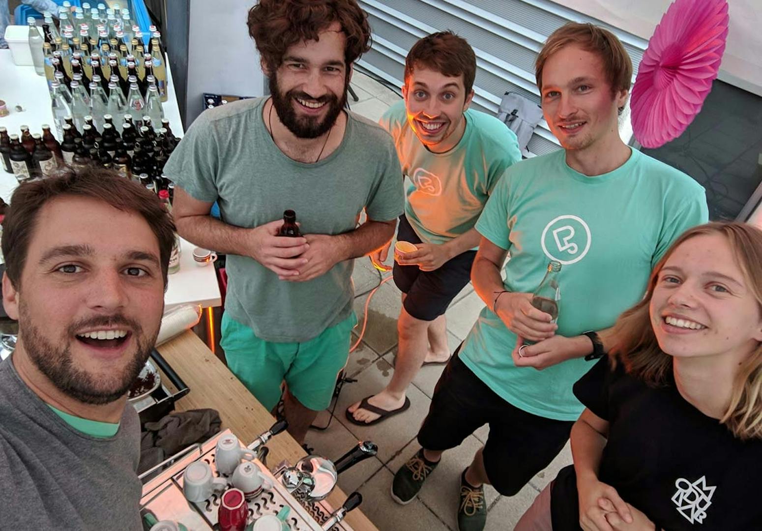 Team photo in the office Five team members in teal and green shirts smiling at the camera in an office with shelves of bottles visible in the background