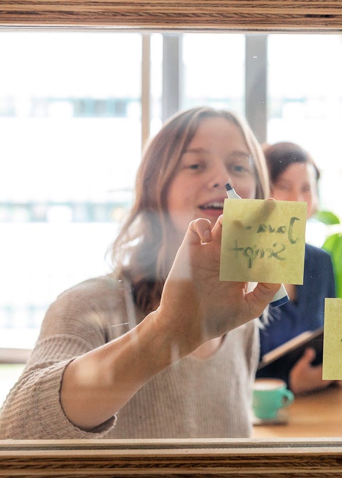 Frau hält gelben Notizzettel mit handschriftlichem Text in die Kamera, lächelt, im Hintergrund weitere Personen in hellem Büro