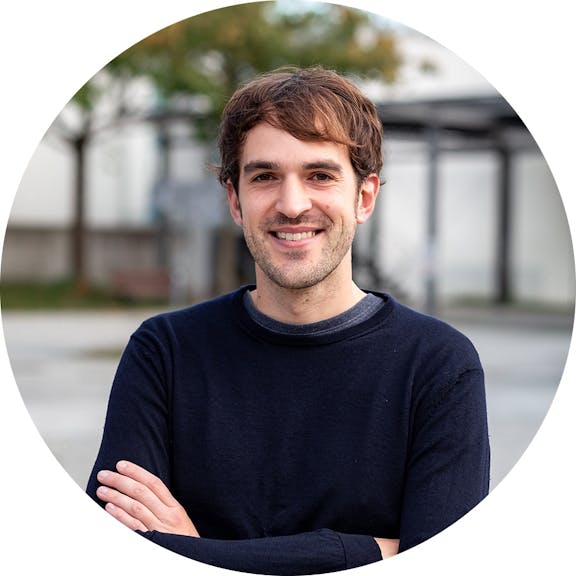 Portrait von Johannes, lächelnder Mann mit braunem Haar in dunklem Pullover vor modernem Gebäude