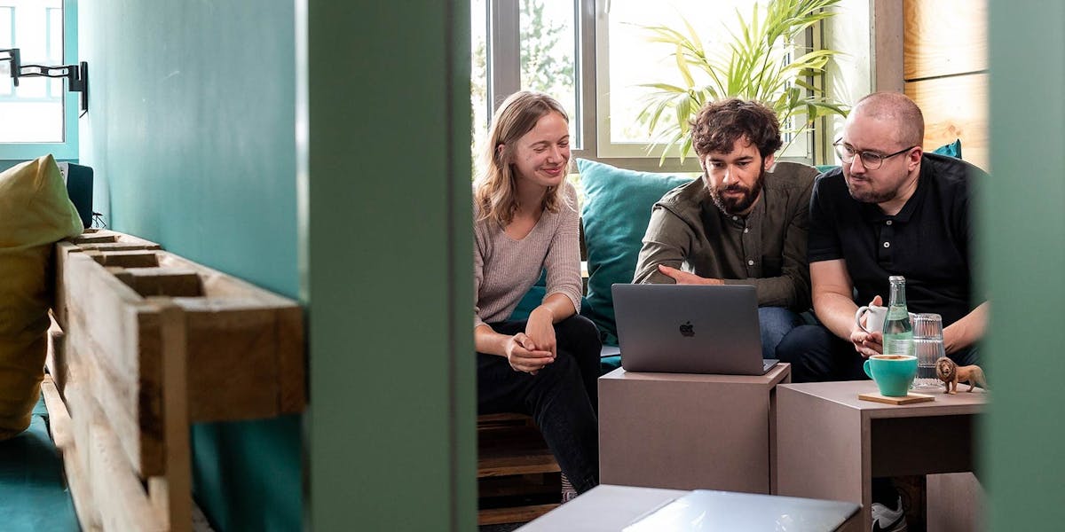 Projektunterstützung im Team Drei Personen sitzen zusammen auf einem Sofa und schauen auf einen Laptop-Bildschirm während einer Projektbesprechung.