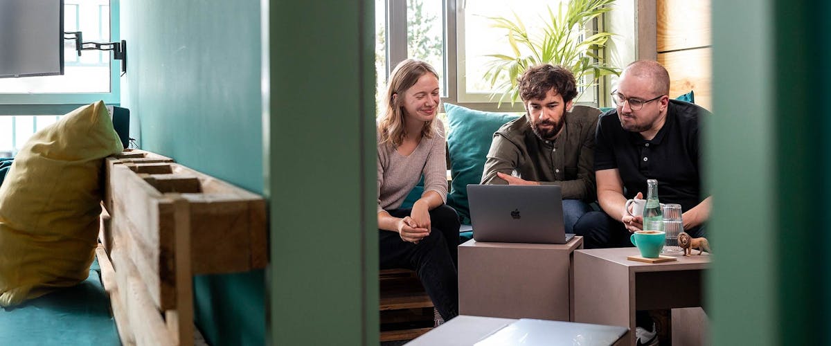 Projektunterstützung im Team Drei Personen sitzen zusammen auf einem Sofa und schauen auf einen Laptop-Bildschirm während einer Projektbesprechung.