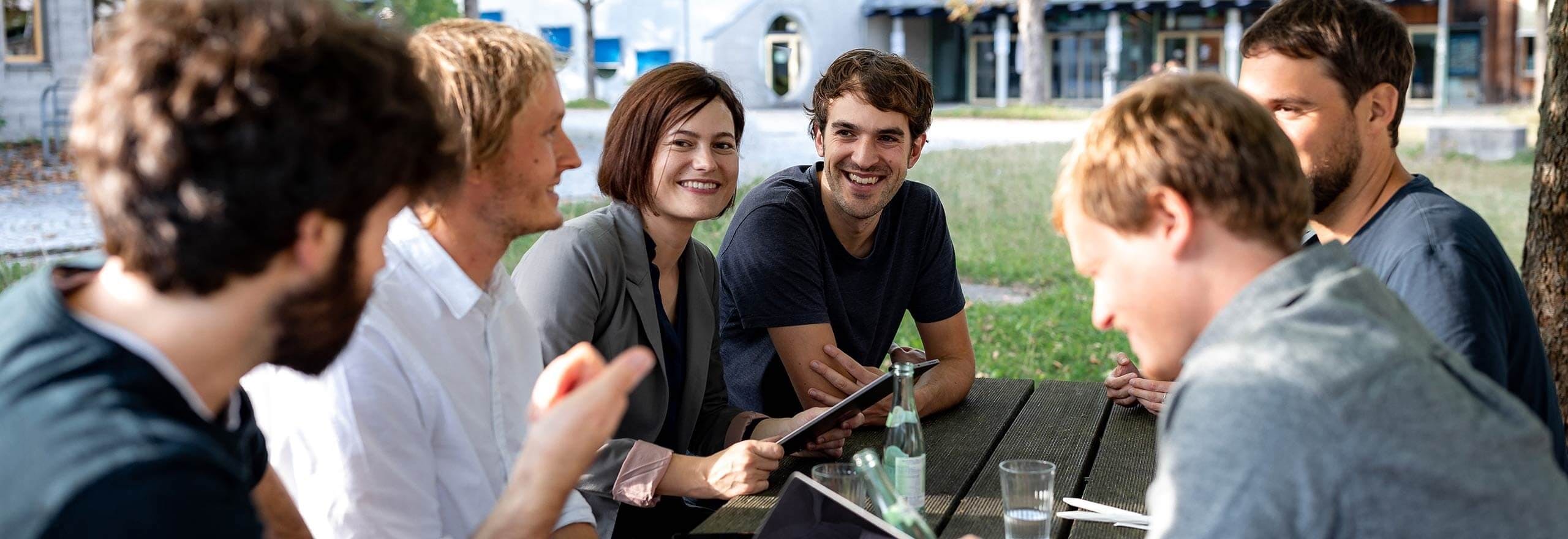 Gruppe von sechs Personen sitzt im Freien zusammen und diskutiert ein Projekt, dabei halten sie ein Tablet und Notizbücher