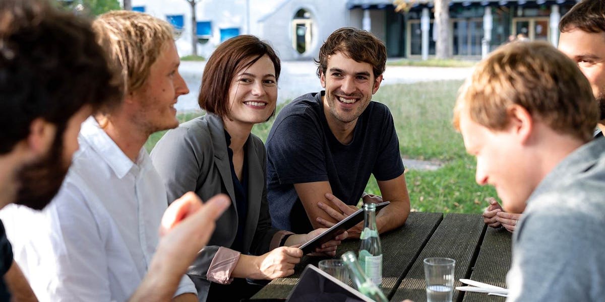 Team-Zusammenarbeit bei der digitalen Produktentwicklung Gruppe von sechs Personen sitzt im Freien zusammen und diskutiert ein Projekt, dabei halten sie ein Tablet und Notizbücher
