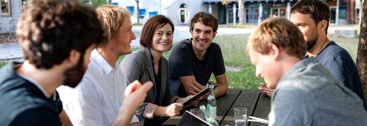 Gruppe von sechs Personen sitzt im Freien zusammen und diskutiert ein Projekt, dabei halten sie ein Tablet und Notizbücher