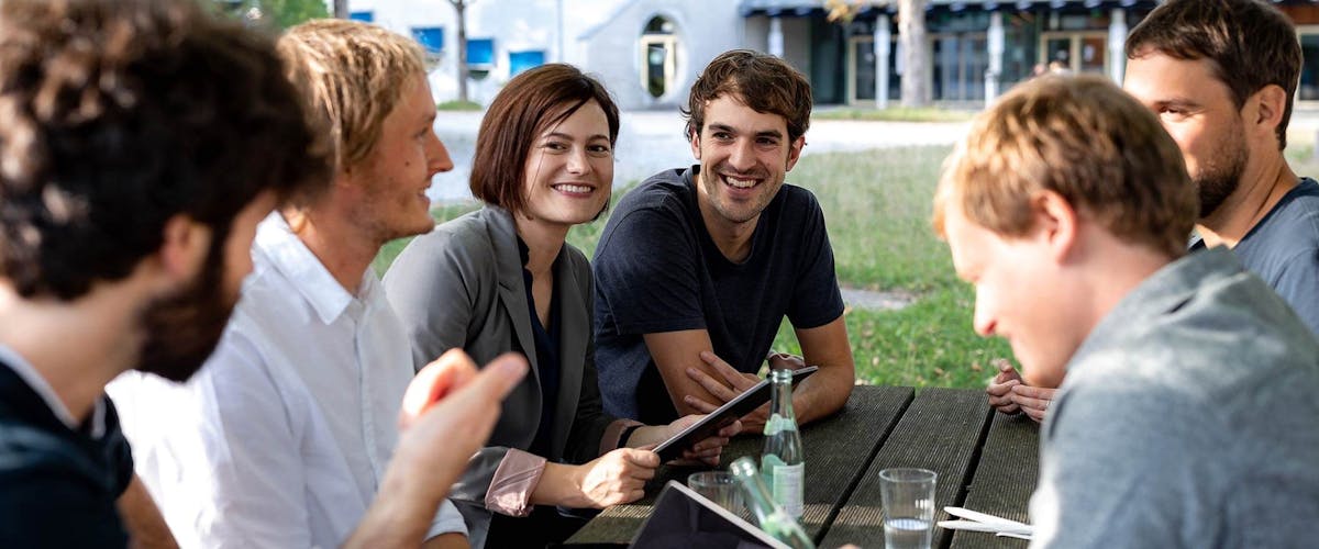 Team-Zusammenarbeit bei der digitalen Produktentwicklung Gruppe von sechs Personen sitzt im Freien zusammen und diskutiert ein Projekt, dabei halten sie ein Tablet und Notizbücher