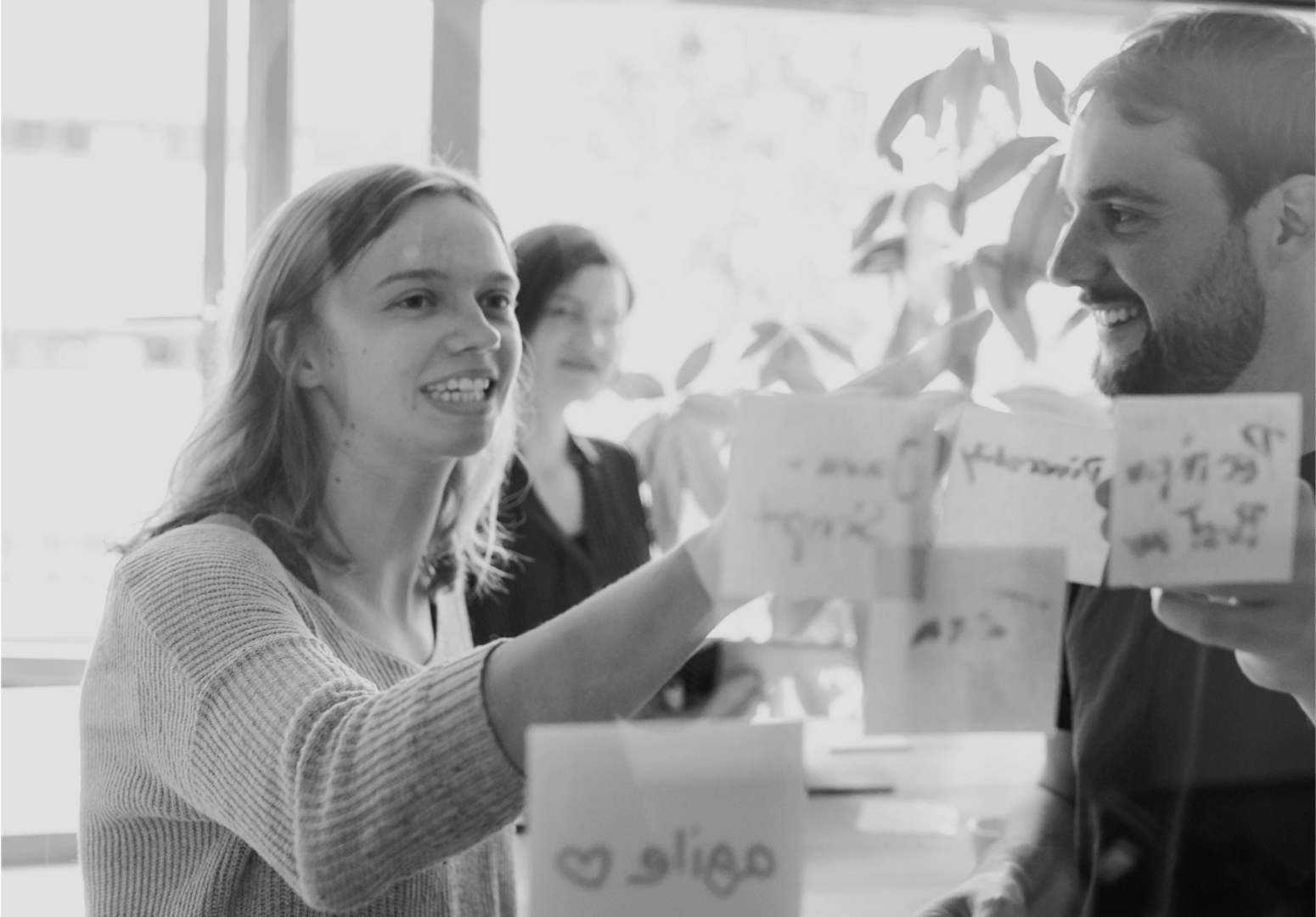 Woman presenting notes on flipchart during workshop, man listening attentively, office environment with plants in background