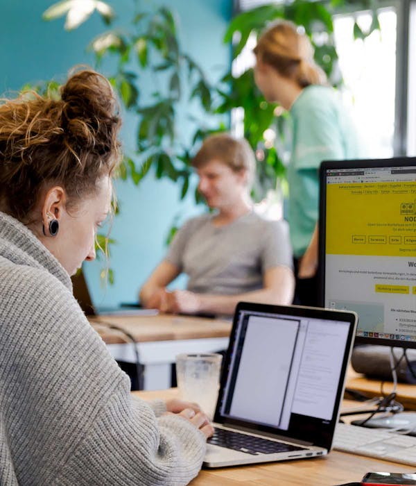 Nodeschool Augsburg: Kostenloses Programmier-Workshop Frau mit Dutt arbeitet konzentriert an Laptop in einem Workshop-Raum mit grünen Pflanzen, im Hintergrund weitere Teilnehmer