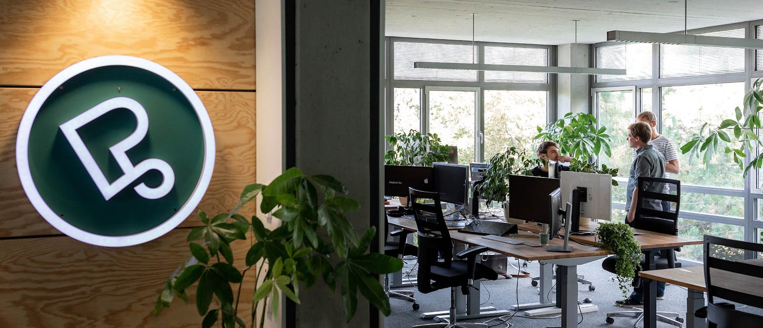 Modernes Büro von Peerigon mit grünem Neon-Logo, Mitarbeitern an Schreibtischen und Pflanzen