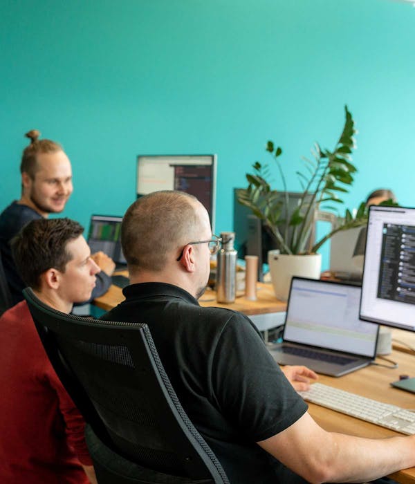 Three developers collaborating at computers in a modern office with turquoise wall and potted plant.
