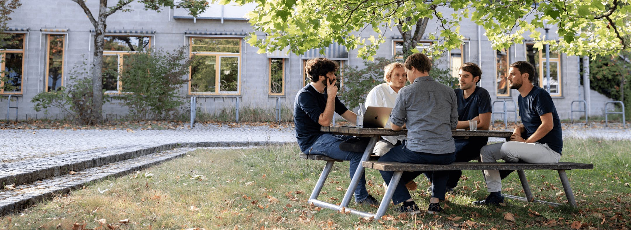 Group of six people sitting at an outdoor table in front of a historic building, collaborating on laptops and documents together.