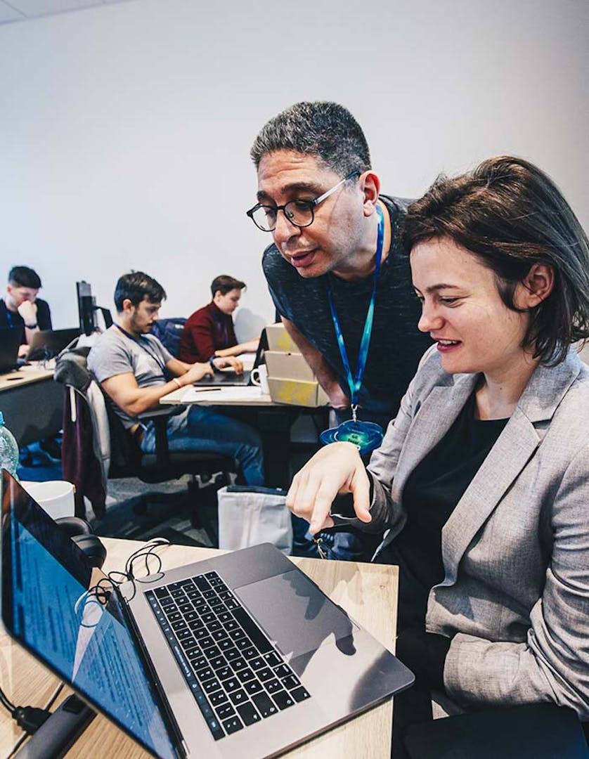 IT-Schulung und Mentoring Trainer mit Brille zeigt einer Frau in grauem Blazer etwas auf einem Laptop in einem Schulungsraum mit anderen Teilnehmern im Hintergrund
