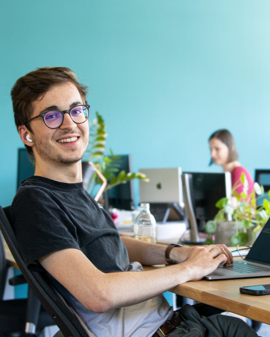 Modernes Arbeitsumfeld mit fokussiertem Team Junger Mann mit Brille sitzt lächelnd am Schreibtisch vor einem Laptop in einem modernen Büro mit hellblauer Wand und Grünpflanzen.