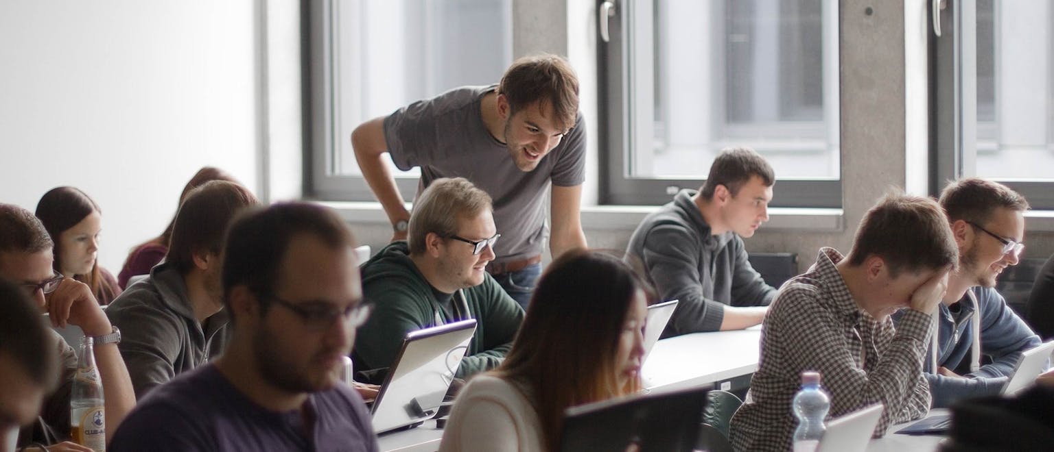Gruppe von Teilnehmern in einem Workshop sitzt an Tischen mit Laptops, ein Mann beugt sich vor und erklärt etwas.