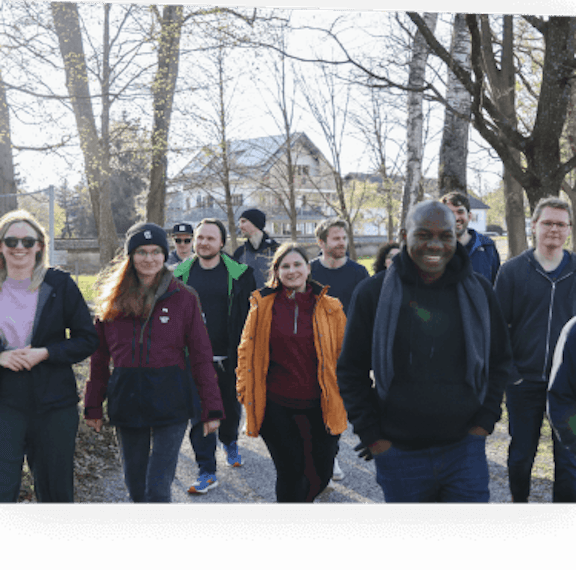 Outdoor Team Group Portrait Group of approximately 15 people of diverse backgrounds standing together smiling on a tree-lined path, with a large house visible in the background.