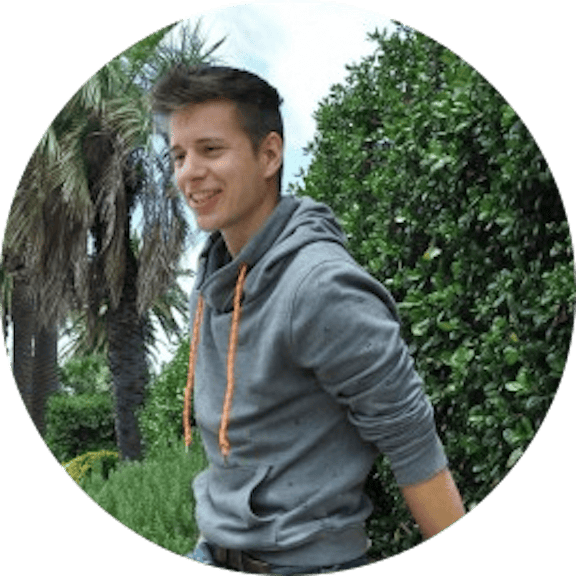 Young man in gray hoodie with orange drawstrings smiling at camera, surrounded by green plants and palm trees